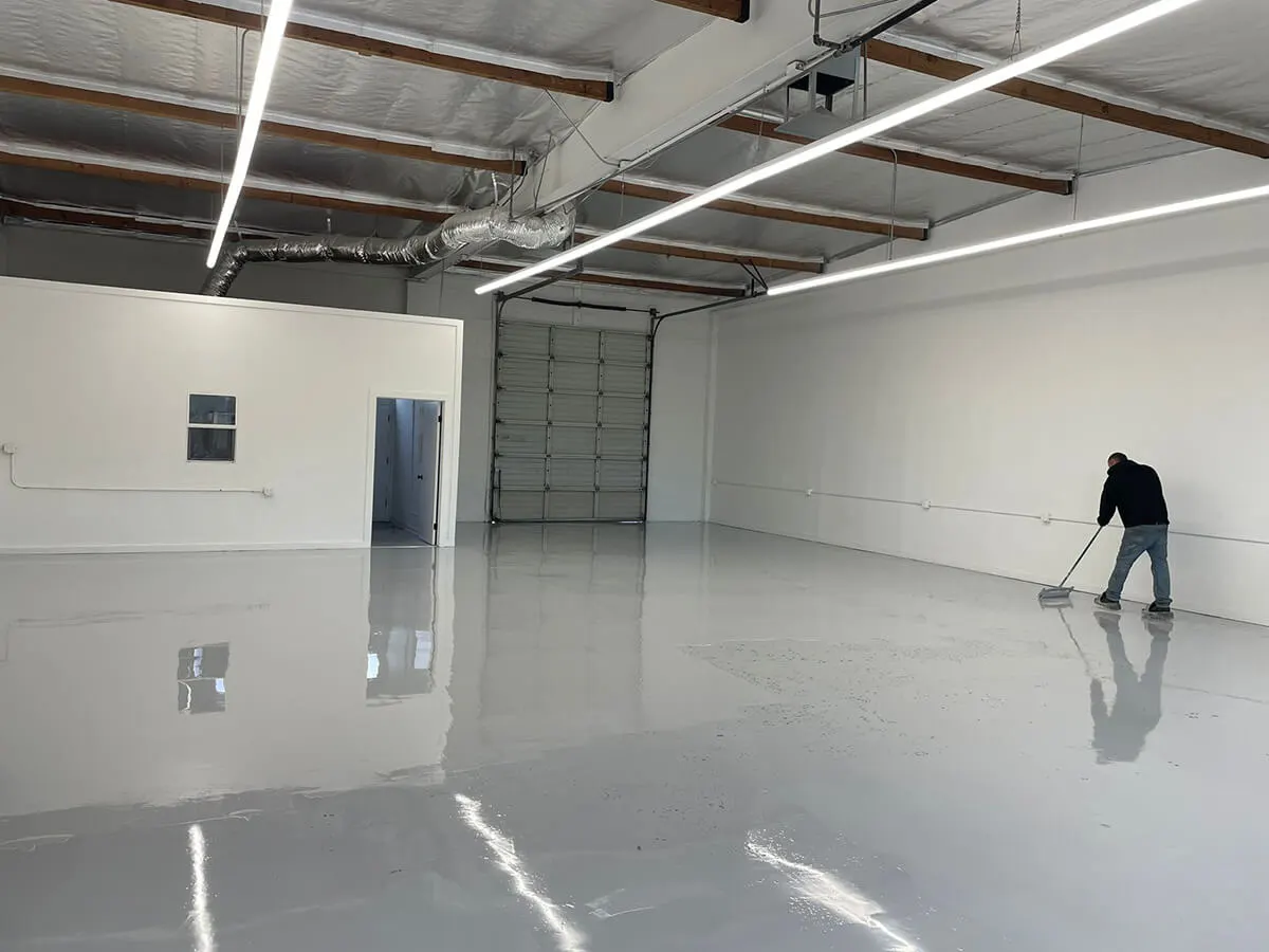 Worker spreading light gray gloss coating across a small warehouse floor